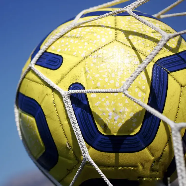 Close-up of a yellow and blue soccer ball caught in the net against a clear blue sky